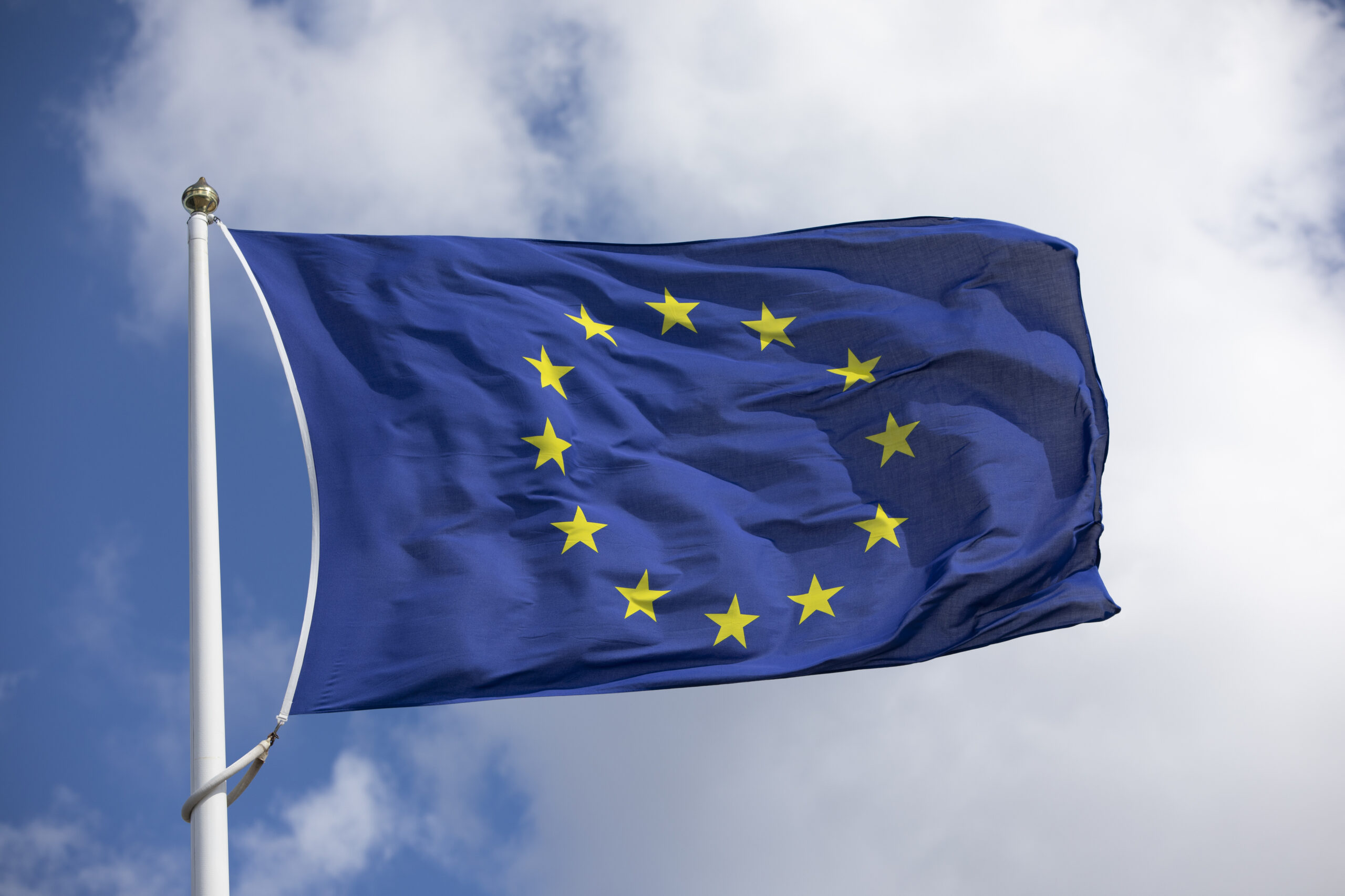 Øverste del av flaggstang med EU-flagget vaiende i vinden mot himmel som er blå med hvite skyer.