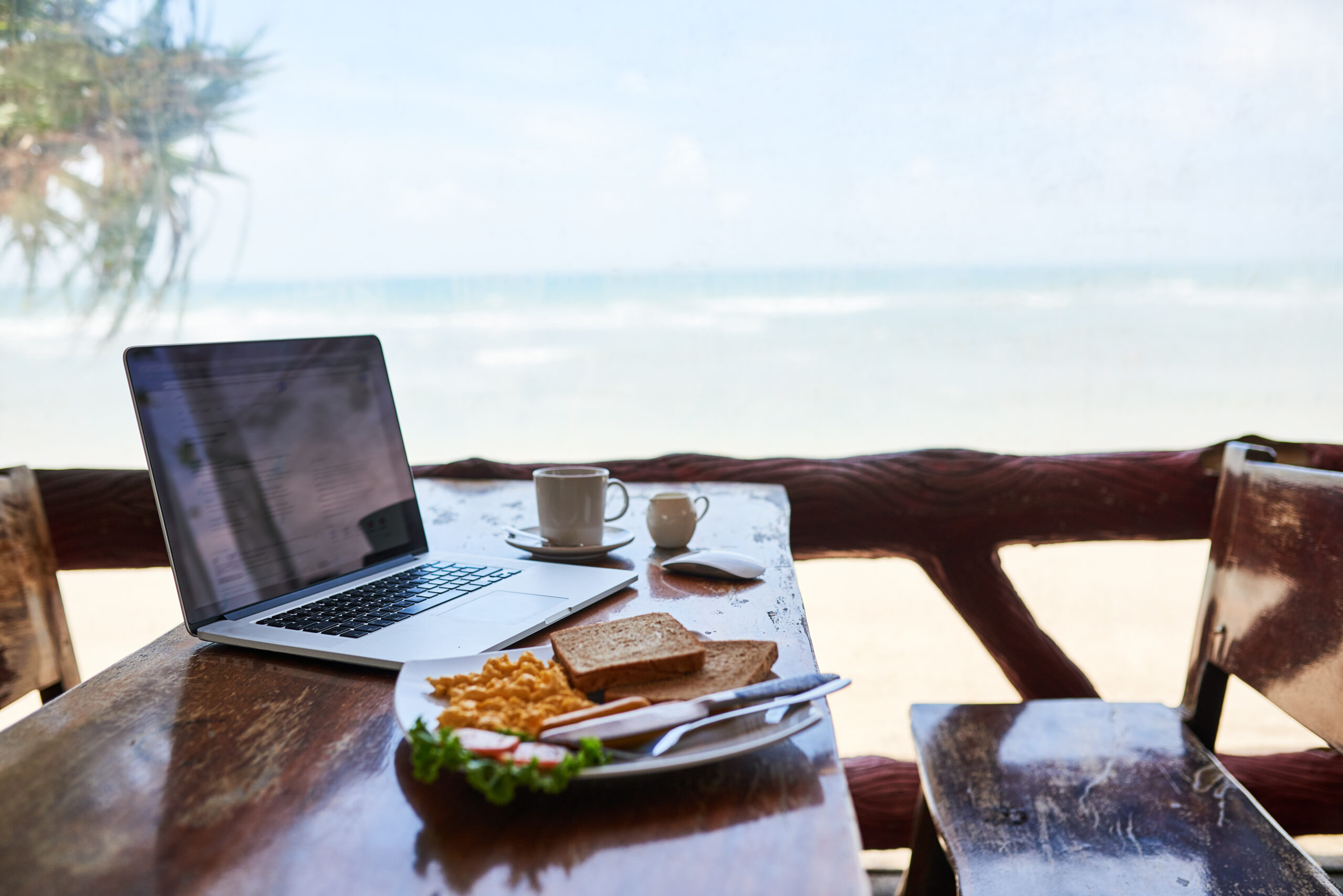 Shot of a laptop and freshly made breakfast on a table with a view of the beach in the background