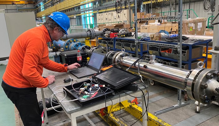 Bildet viser en person som står ved en pc i et industrimiljø med rørgate og arbeider med massestrømsmåling av CO2, her ved vannstrømstest.