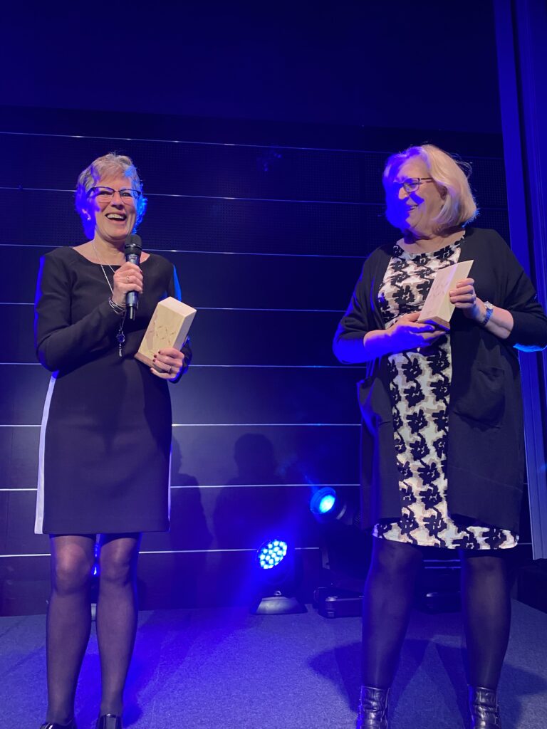 Two happy women in dresses standing on a stage in bluish light. One is holding a microphone, both are holding the award/figure they have received. Photo