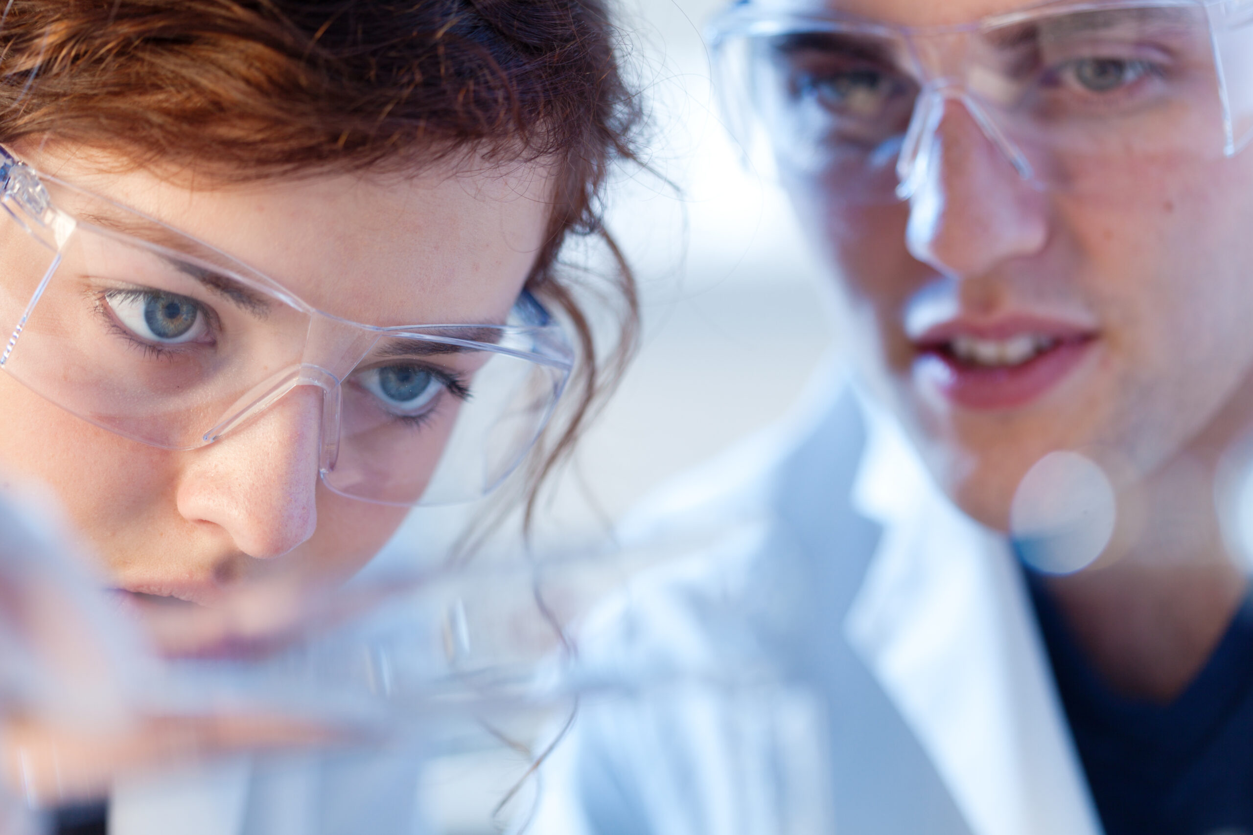 To forskere i et kjemilaboratorium, en ung mann og en ung kvinne, arbeider sammen i et forskningslaboratorium for biologi og kjemi på et universitet. Fokus på laboratorieutstyret med personalet i bakgrunnen. Fotografert i horisontalt format.